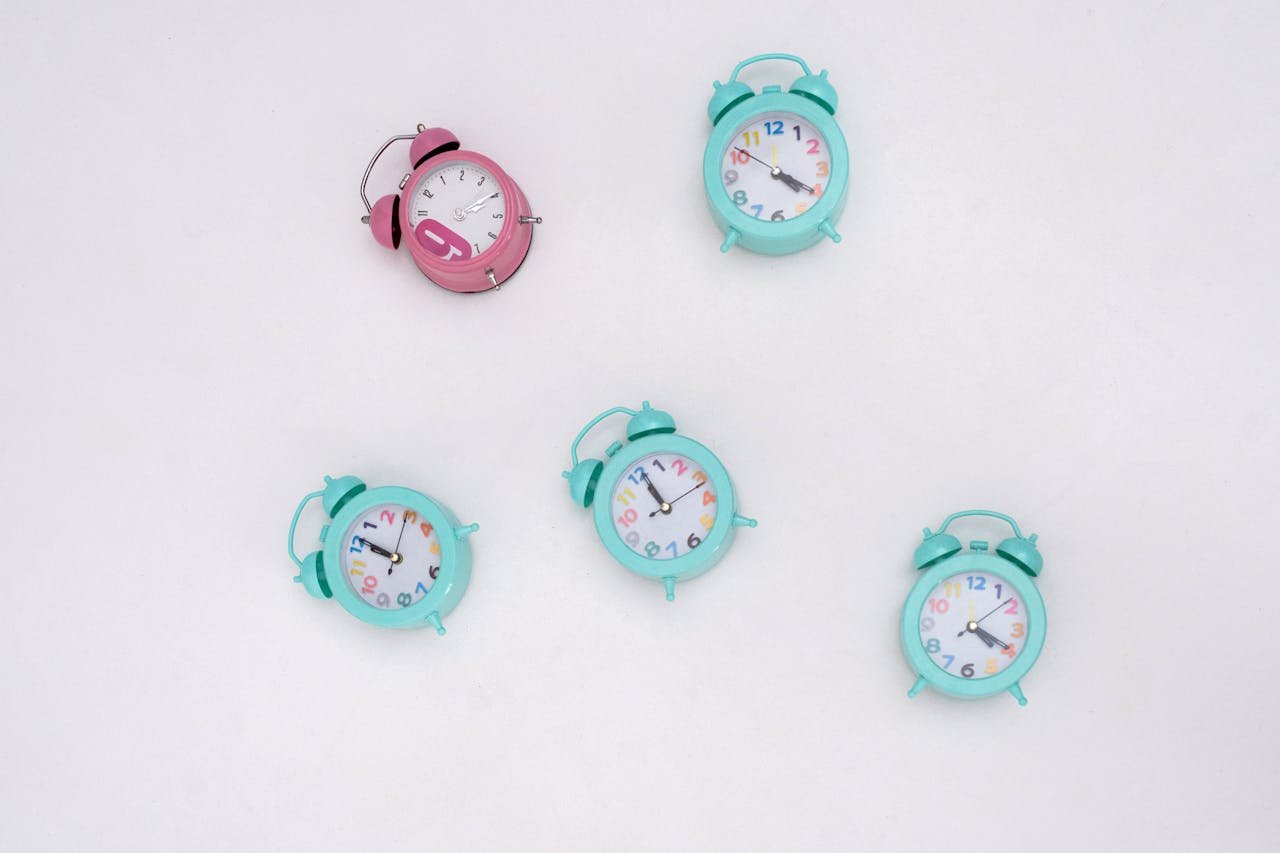 Five colorful analog alarm clocks arranged on a white background in a minimalist setup.